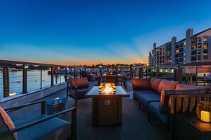A rooftop bar with outdoor seating, a fire pit, and a view of the marina at dusk.