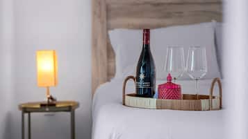 Una cama bien hecha con una botella de vino rosado, dos copas y un recipiente decorativo sobre una bandeja de mimbre.