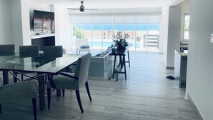 A spacious living area with a dining table, chairs, a TV, and a view of the ocean through large windows.