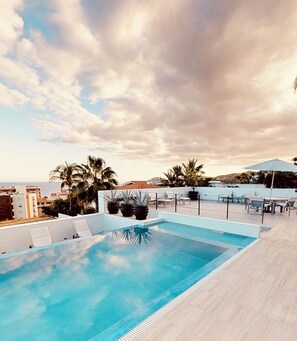 A rooftop pool with lounge chairs and umbrellas, surrounded by palm trees and buildings.