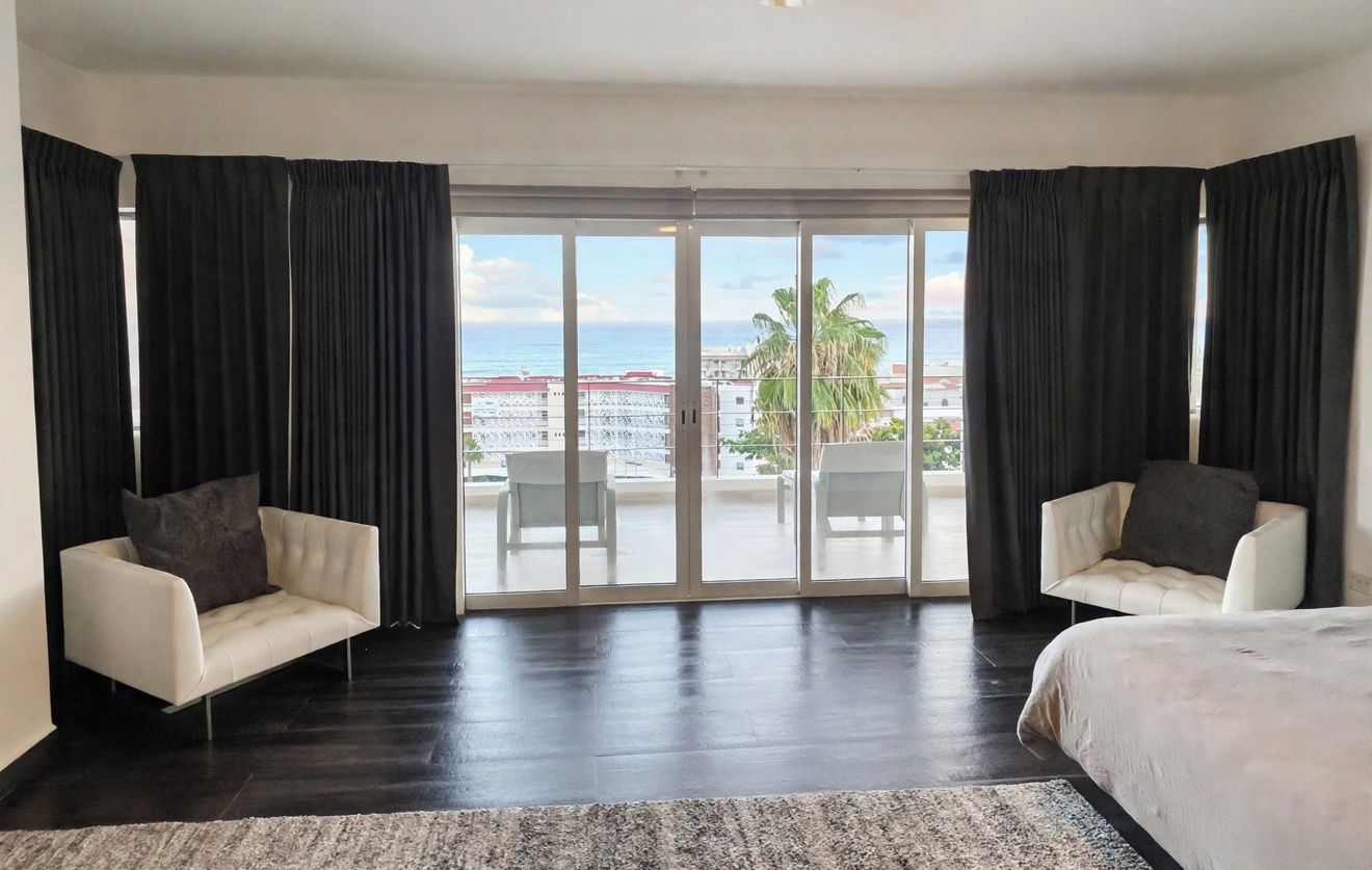 A hotel room with a large sliding glass door leading to a balcony, two armchairs, a bed, and a dark wooden floor.