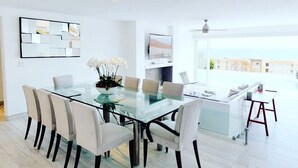A modern dining area with a glass table and white chairs, a TV mounted on the wall, and a view of the sea through large windows.