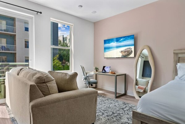 A modern bedroom with a large bed, a desk with a laptop, a chair, and a window with a view of a building and greenery. - Channelside: Workers Paradise| Near Everything (Tampa)