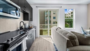 A modern kitchen with dark cabinets, a black countertop, and a stainless steel range hood. There is a sliding glass door leading to a balcony with a view of a building and greenery. A gray sofa is positioned near the window. - Channelside: Workers Paradise| Near Everything (Tampa)