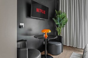 A modern living room with a flat-screen TV displaying 'NETFLIX,' a round table with an orange lamp, two black leather chairs, and a potted plant. - Trendy Ybor Studio | Free Parking | Close to DT! (Tampa)