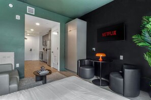 Ein modernes Hotelzimmer mit Bett, kleinem Tisch, Wand-Fernseher und Blick in den Küchenbereich.