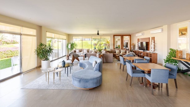 A spacious living area with a large sofa, dining table, and chairs, a kitchen area with cabinets and a refrigerator, and large windows offering a view of a garden.