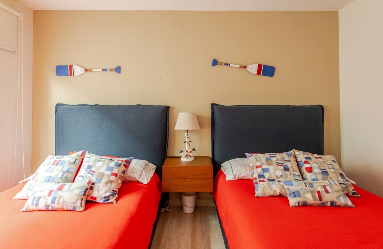 A hotel room with two beds, each with red bedding and pillows with a pattern. There is a nightstand with a lamp between the beds.