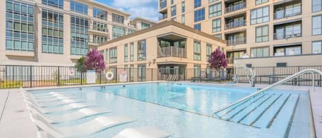 Una piscina con sillas de descanso, rodeada por un edificio con varios balcones y amplias ventanas.
