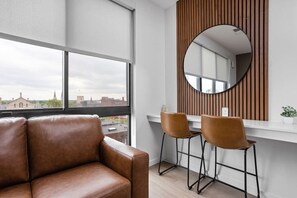 A modern room with a large window, a leather sofa, two bar stools, and a round mirror on a wooden panel wall.