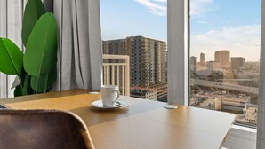 A hotel room with a large window offering a city view, a wooden table with a cup, and a potted plant.