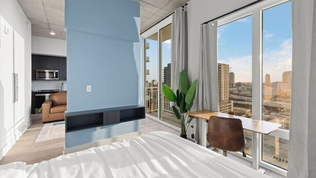 A modern hotel room with a large bed, a desk with a chair, a view of the city, and a kitchenette in the corner.