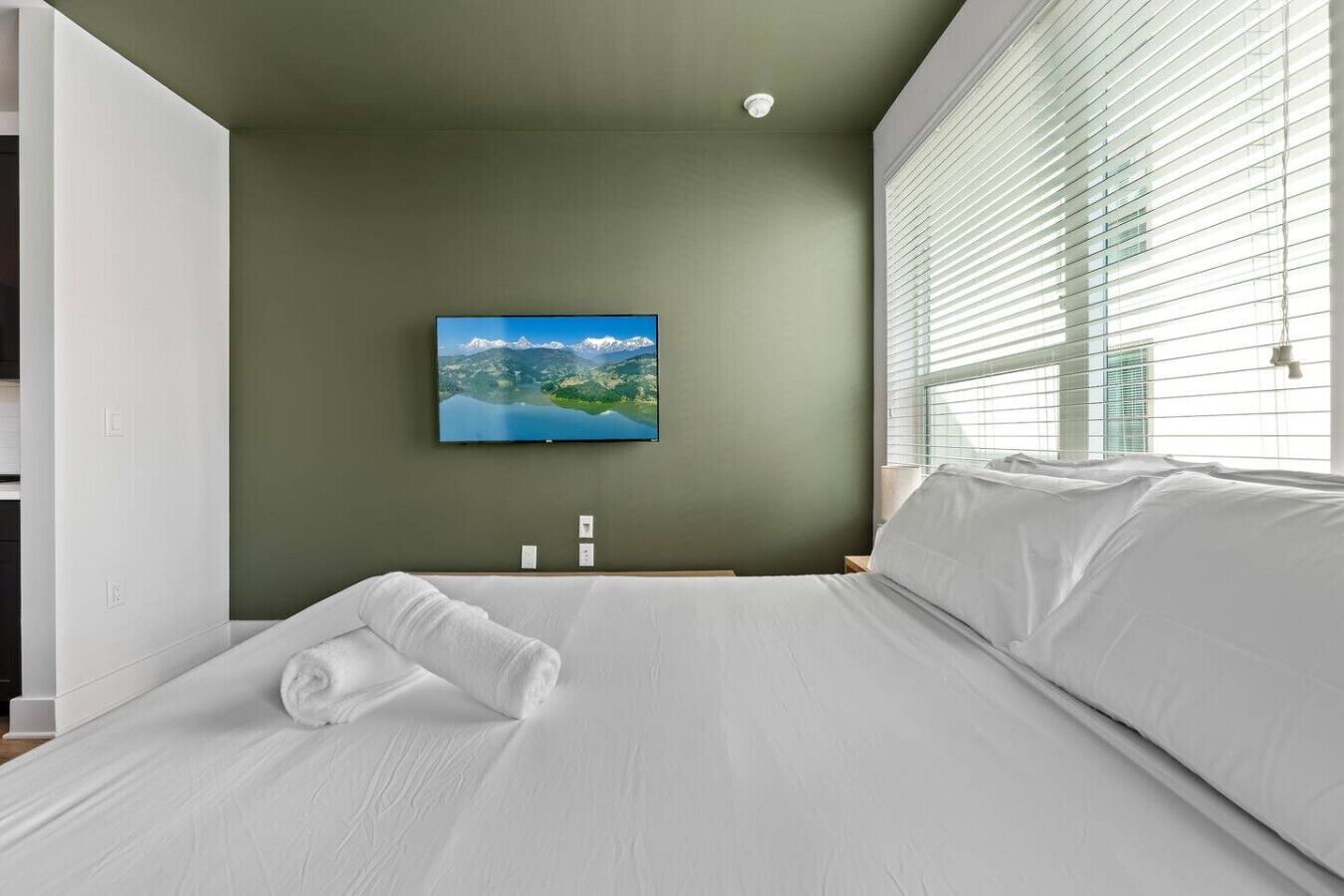 Habitación de hotel con cama, televisor de pantalla plana en la pared y ventana con persianas.