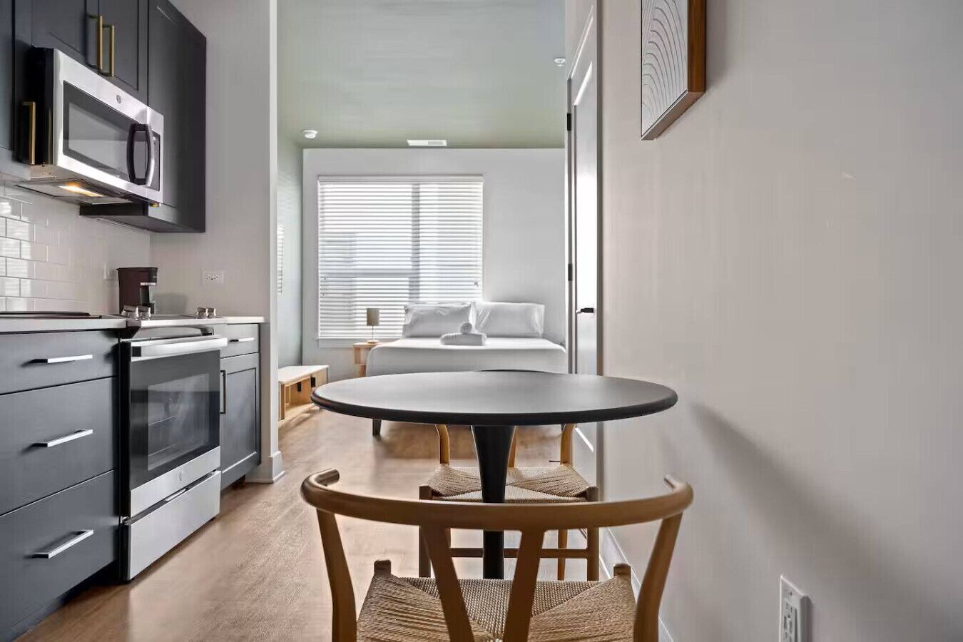 Una cocina compacta con electrodomésticos modernos, un comedor con una mesa redonda y una silla de madera, y un dormitorio con cama y ventana.