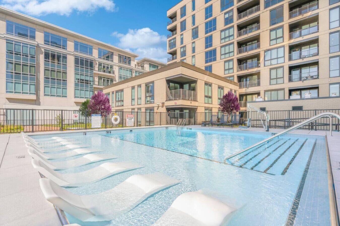 A swimming pool with lounge chairs, a building with multiple floors, and a clear sky.