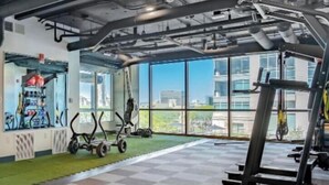 A gym with exercise equipment, large windows, and a view of buildings. - Comfy Downtown Studio| Pool/Parking/Gym (Indianapolis)