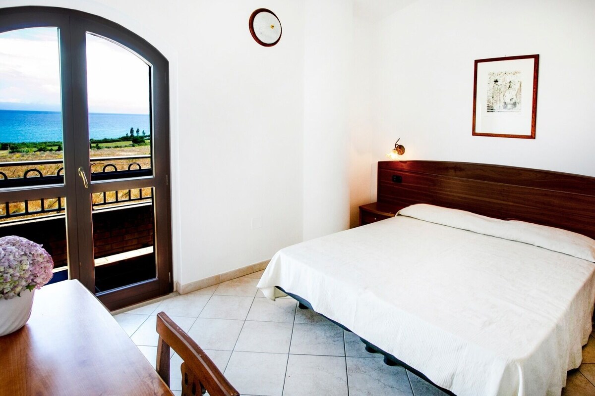 A bedroom with a bed, a bedside table, a chair, a clock, and a framed picture on the wall.