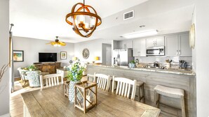 Cocina moderna con armarios blancos, encimera de mármol y electrodomésticos de acero inoxidable. Cuenta con una zona de comedor con mesa y sillas de madera, y un ventilador de techo.