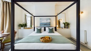 Una cama con dosel, un colchón blanco, dos almohadas, una manta verde y una cesta de frutas.