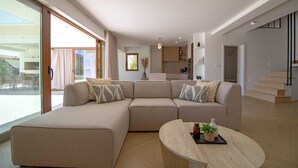 A modern living room with a sectional sofa, a round coffee table, and a view of the kitchen and outdoor area. - Villa Mood – Pool, Gym & Sauna near Split (Kastel Luksic)