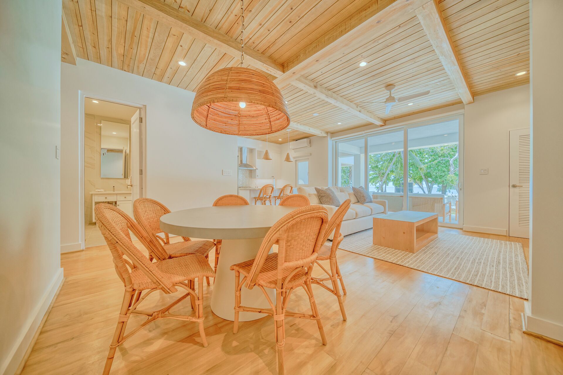 A spacious living area with a round table surrounded by wicker chairs, a large woven light fixture, and a view of the outdoors through large windows.