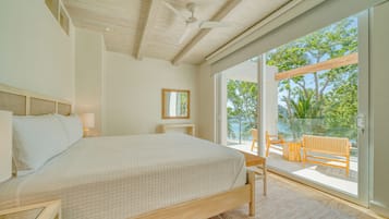 A bedroom with a large bed, a ceiling fan, a window with a view of trees and a beach, and a small table with a chair.