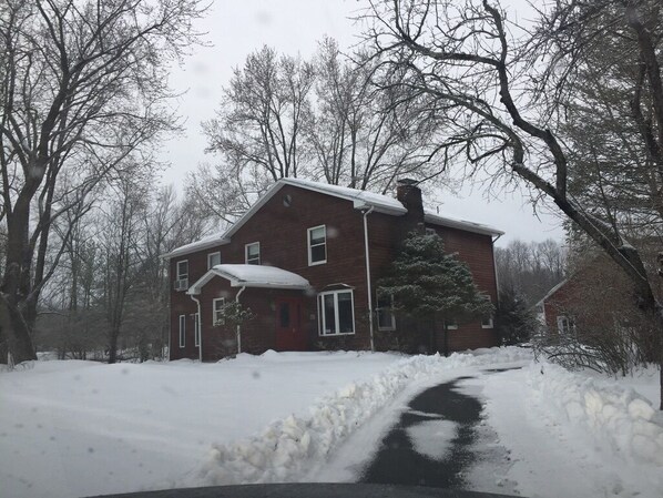 A two-story brick house with a snow-covered driveway and surrounding trees.