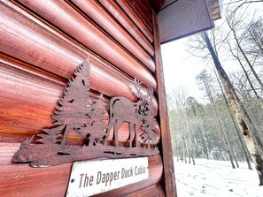 Eine rustikale Holzhütte mit einer Holzfassade, einem Schild mit der Aufschrift 'The Dapper Duck Cabin' und einem verschneiten Wald im Hintergrund.