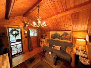 A cozy cabin interior with a wooden ceiling, a chandelier, a sofa with animal-themed pillows, a fireplace, and a window with a wreath.