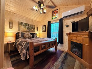 A wooden cabin bedroom with a bed, a fireplace, a TV, and a window with curtains.