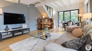 Een moderne woonkamer met een flatscreen televisie, een glazen salontafel, een bank met gekleurde kussens en een trap naar een eethoek.