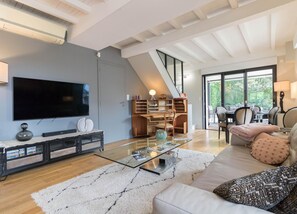 Een moderne woonkamer met een flatscreen televisie, een glazen salontafel, een bank met gekleurde kussens en een trap naar een eethoek.