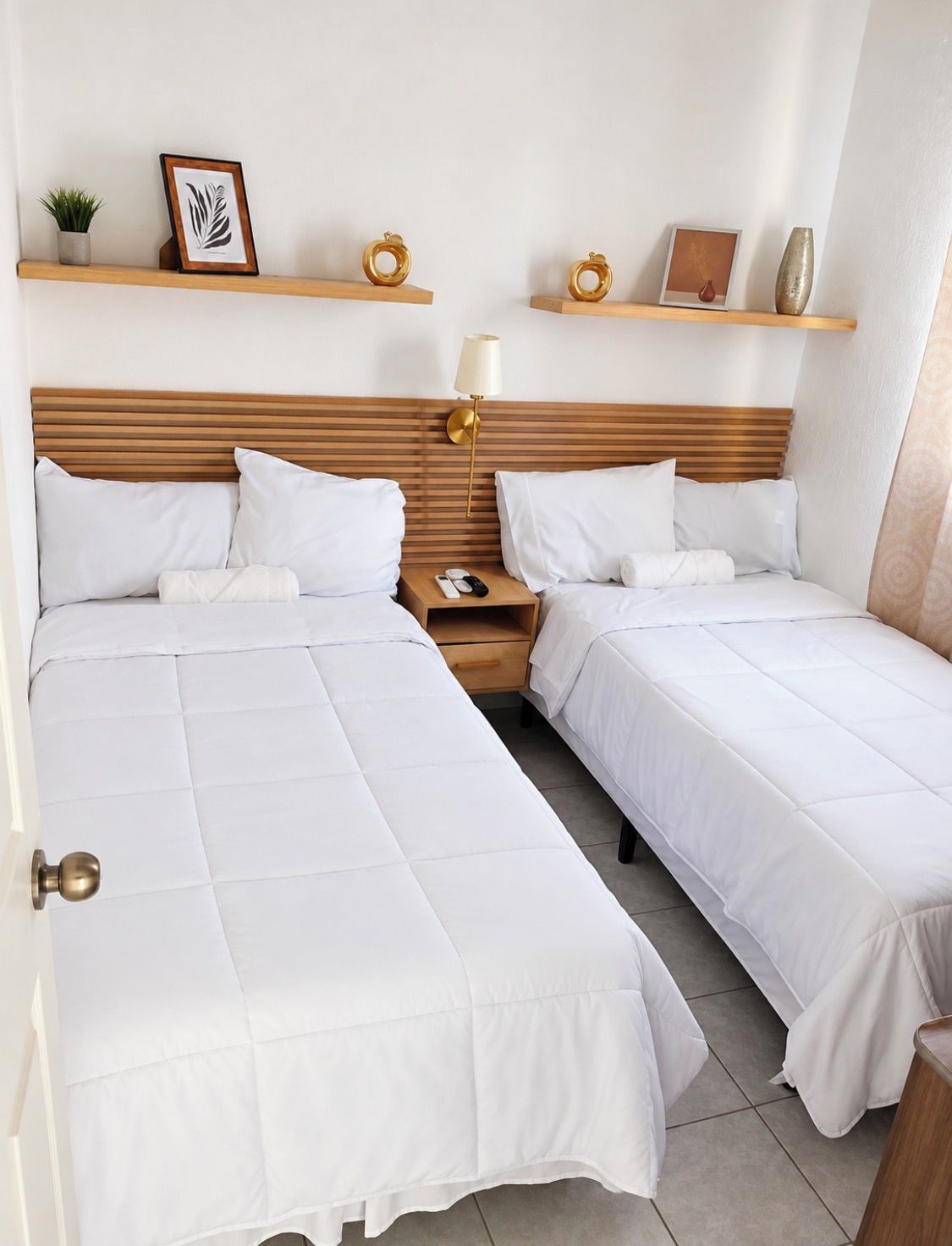 A hotel room with two single beds, a wooden headboard, bedside tables with lamps, and decorative items on shelves.