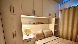 Quarto com cama, mesa de cabeceira, prateleira com objetos decorativos e guarda-roupa.
