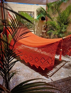 Un hamac avec un auvent rouge, installé dans une cour dont le sol est recouvert de carreaux et où l’on trouve des plantes en pot.