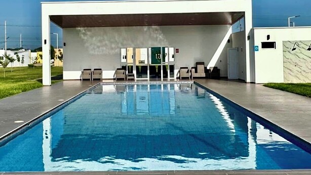 Piscina moderna ao ar livre, com céu azul límpido, deck de tijolos e área coberta com cadeiras.