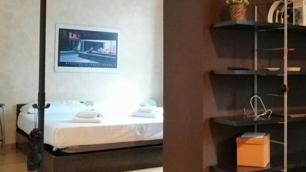 Una habitación de hotel con una cama, un escritorio, una silla, una estantería con objetos y un cuadro en la pared.