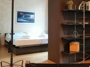 Una habitación de hotel con una cama, un escritorio, una silla, una estantería con objetos y un cuadro en la pared.