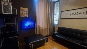 Una habitación con un sofá de cuero negro, un pequeño taburete negro, un televisor sobre un soporte, una obra de caligrafía enmarcada en la pared y una ventana con cortinas.