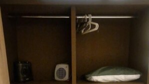 Una habitación compacta con una cama, un pequeño rincón de descanso con una silla, una estantería con un ventilador y un cesto de basura.