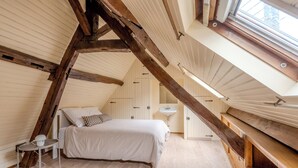 A bedroom with a sloped ceiling, wooden beams, a bed, a bedside table, and a window.