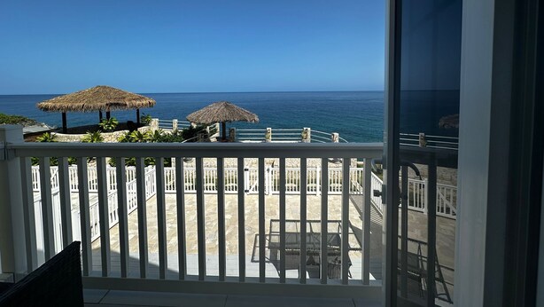 A balcony with a view of the ocean, thatched umbrellas, and a stone wall. - Ocean View Villa Tree House @ Little Bay (Hanover Parish)