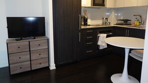 A modern kitchen with dark cabinetry, a built-in oven, a microwave, and a coffee maker. There is a flat-screen TV on a wooden stand, a small table with a white chair, and a towel hanging on the oven door. - Studio + Balcony | Nice DT Condo beside Queen St W (Toronto)