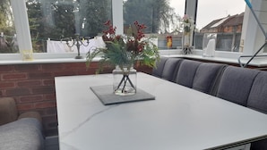 Un espace repas moderne avec une table en marbre blanc, des bancs recouverts d’un tissu gris et un vase en verre contenant des plantes vertes.