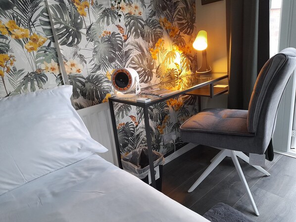 A hotel room with a bed, a chair, a desk with a lamp, and a wall with tropical wallpaper.