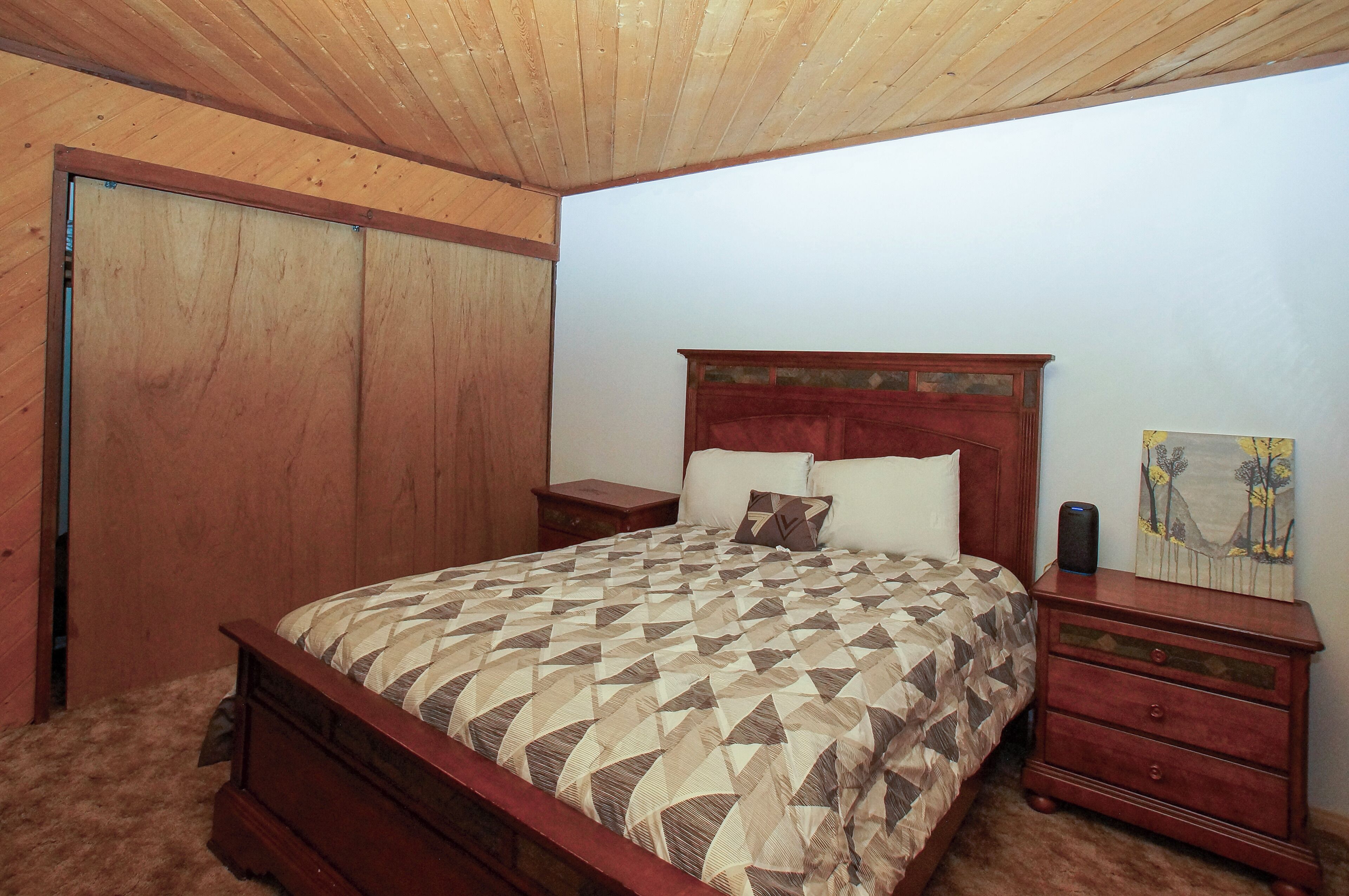 A bedroom with a wooden ceiling, a bed with a patterned bedspread, a nightstand, a dresser, and a painting on the wall.