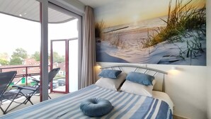 Ein Schlafzimmer mit Bett, Kissen und Blick auf einen Strand durch ein großes Fenster.