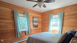 A bedroom with wooden walls, a ceiling fan, a bed with patterned bedding, a bedside table with a lamp, and a rug on the floor.