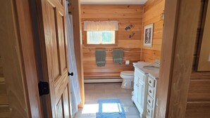 A wooden cabin bathroom with a window, a toilet, a sink, and a shower area.
