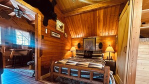 A wooden cabin bedroom with a bed, two lamps, a ceiling fan, and a mounted bear head. - The Raven tiny log cabin in The Forest (Cosby)